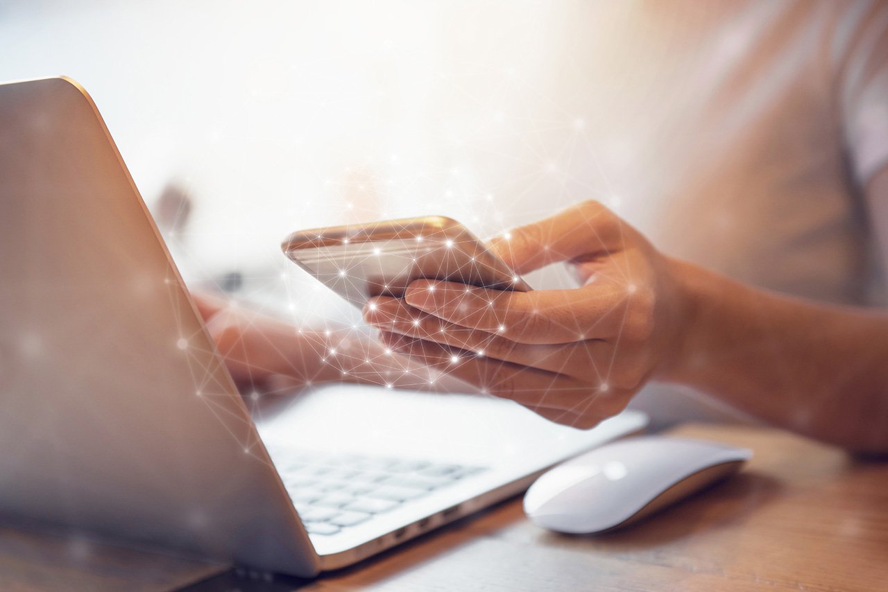 close up image of hands holding a cellphone and using a laptop